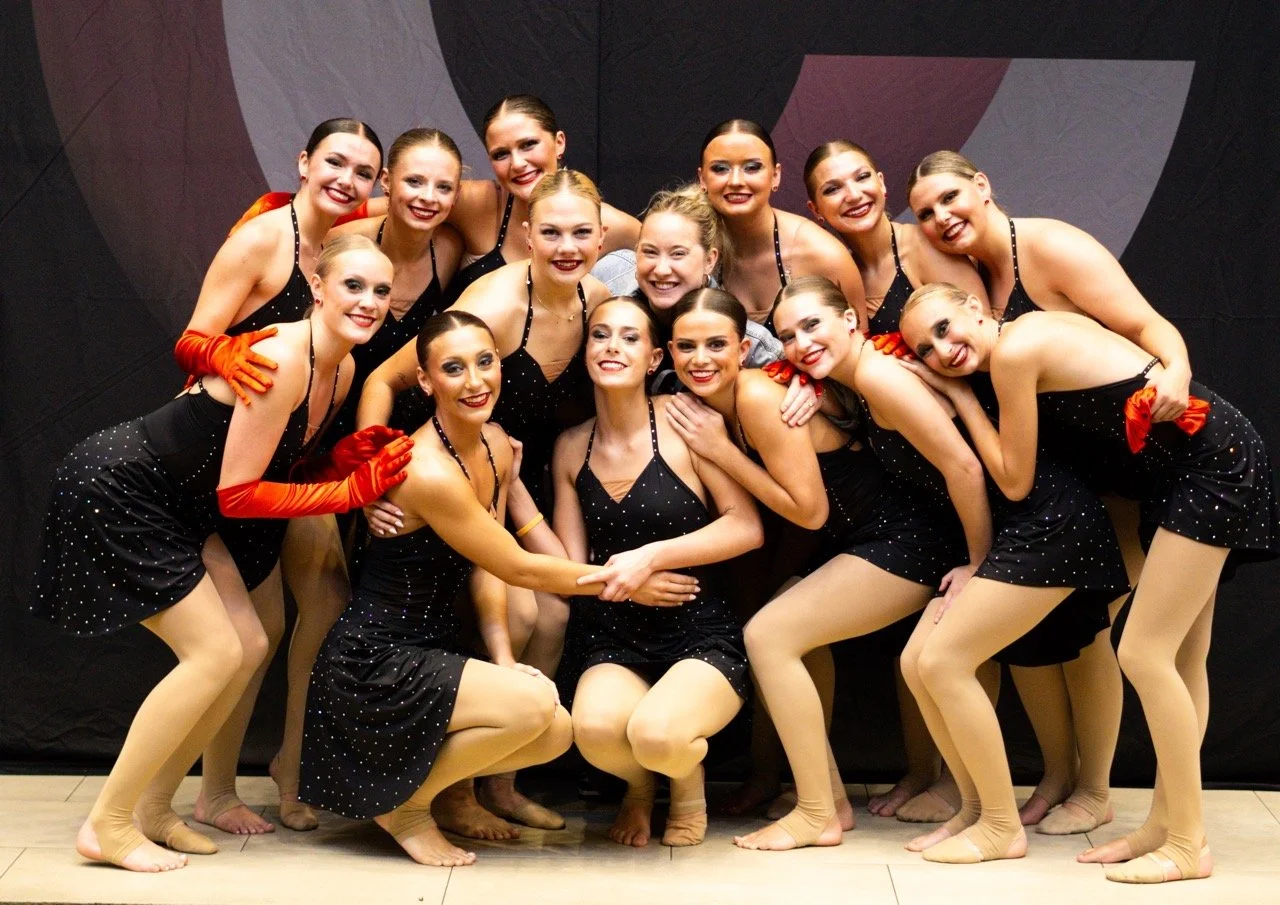 A group of fifteen young female dancers posing together backstage, wearing black polka-dotted costumes with beige tights and some with orange gloves, smiling at the camera.