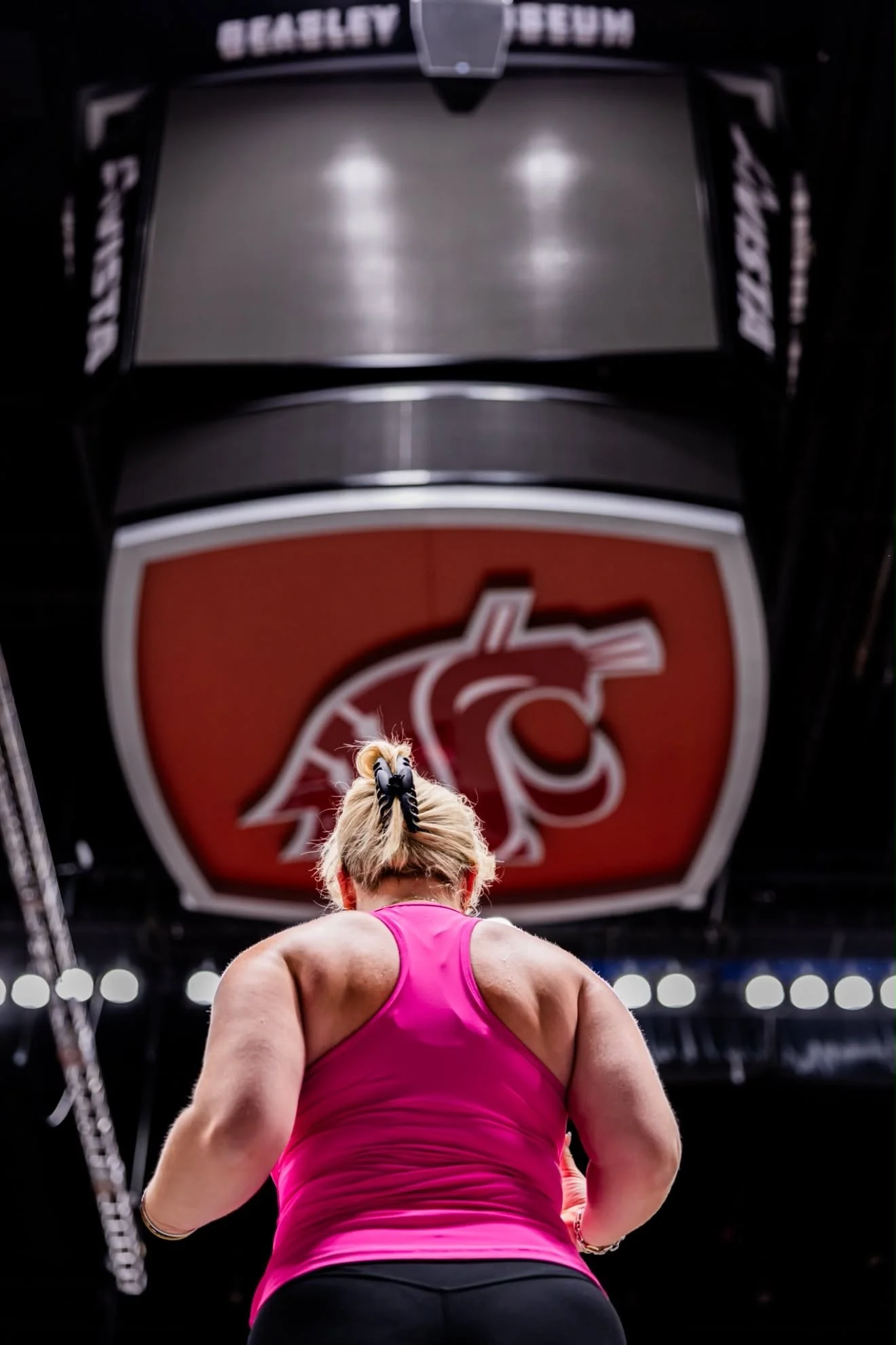 Female athlete with blond hair in a bun, wearing a pink tank top, seen from the back, preparing for a workout in a gym with a large Kansas State Wildcats logo on the ceiling.