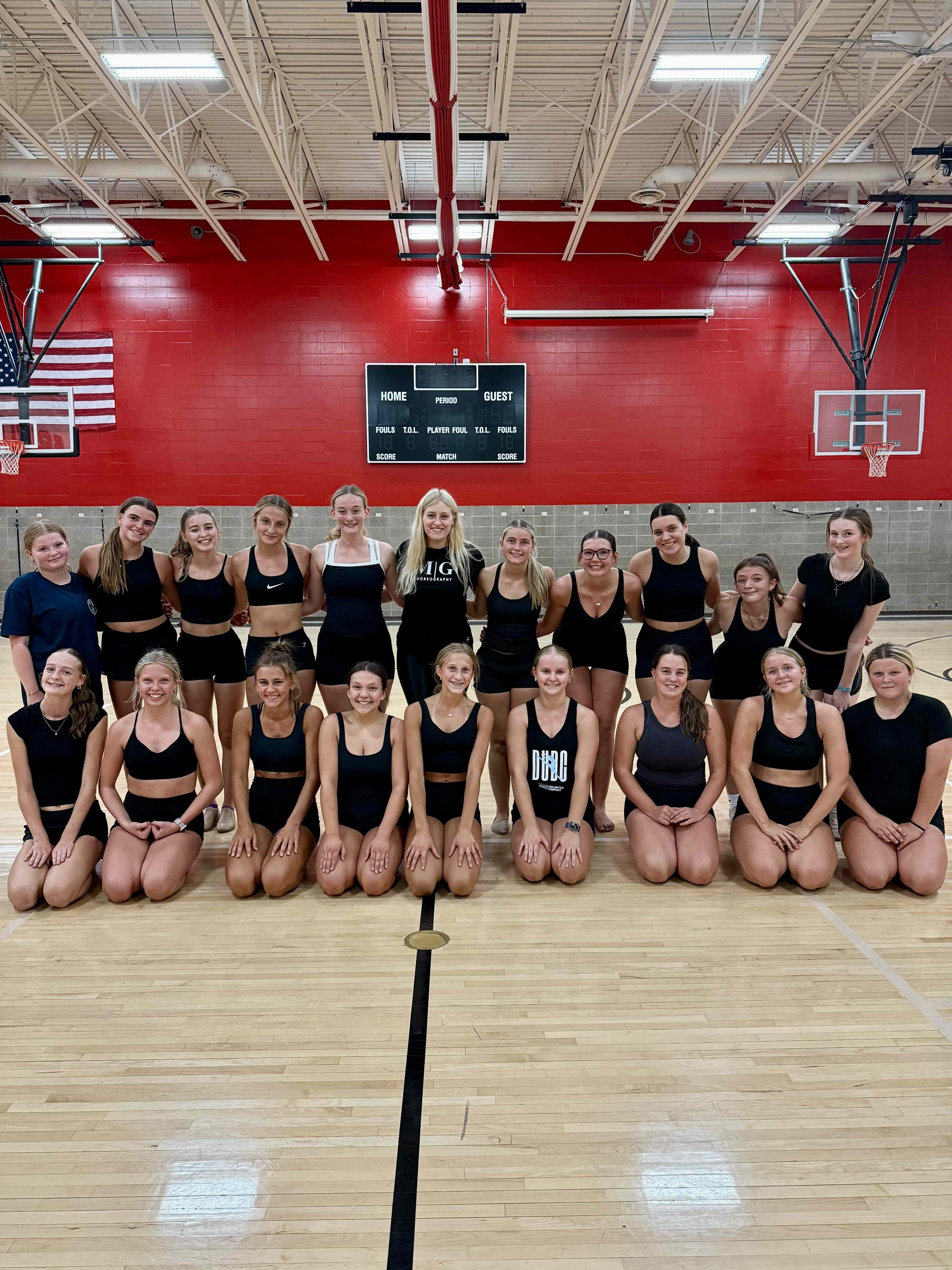 A group of young female cheerleaders posing for a group photo on a basketball court with a red wall, basketball hoops, and scoreboard in the background.