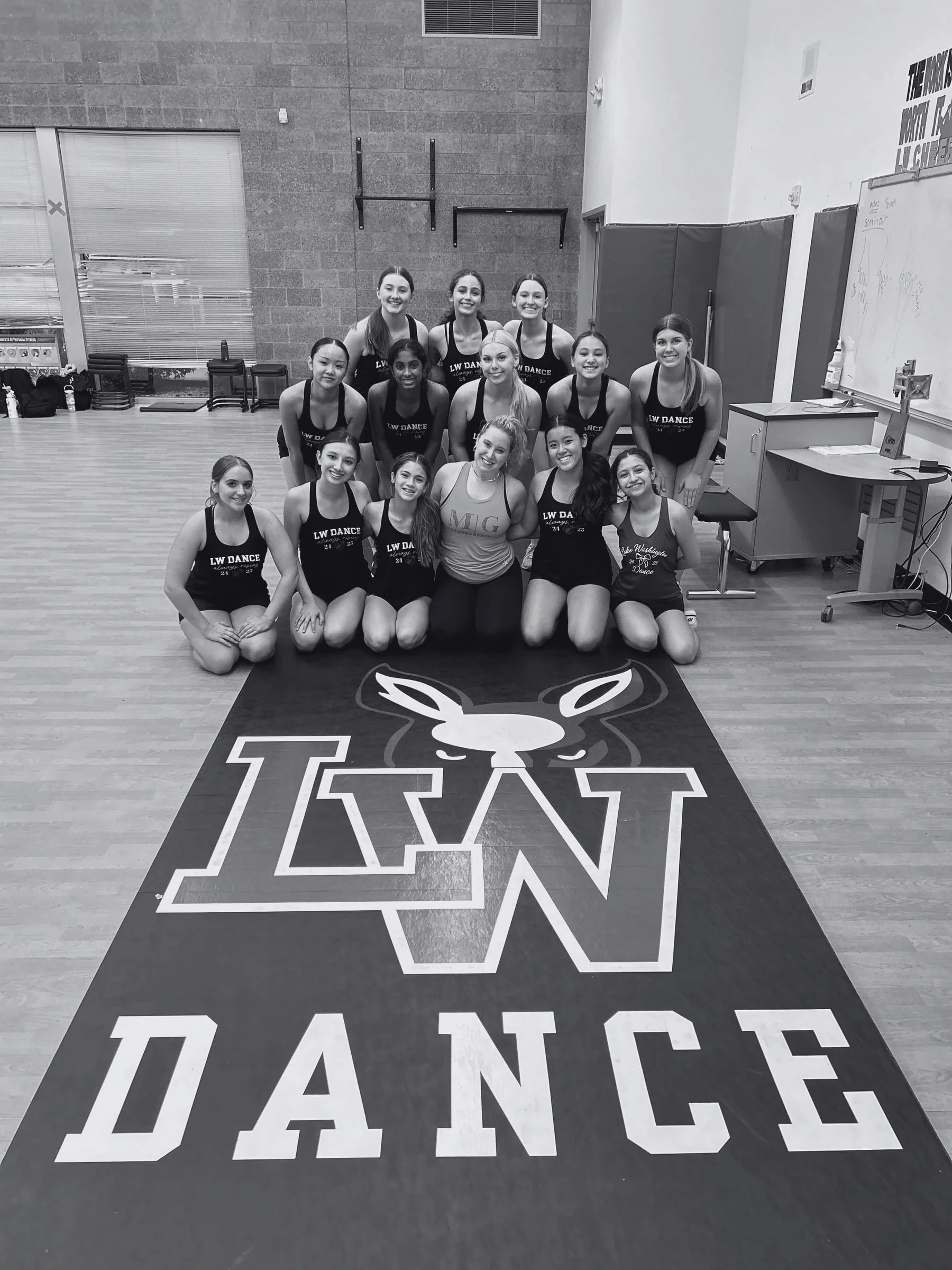 A group of 13 young women posing for a photo in a dance studio, standing on a floor mat with the word 'DANCE' and a bunny logo. The women are wearing matching black dance uniforms with 'LW DANCE' printed on them. The room has a brick wall, large wind
