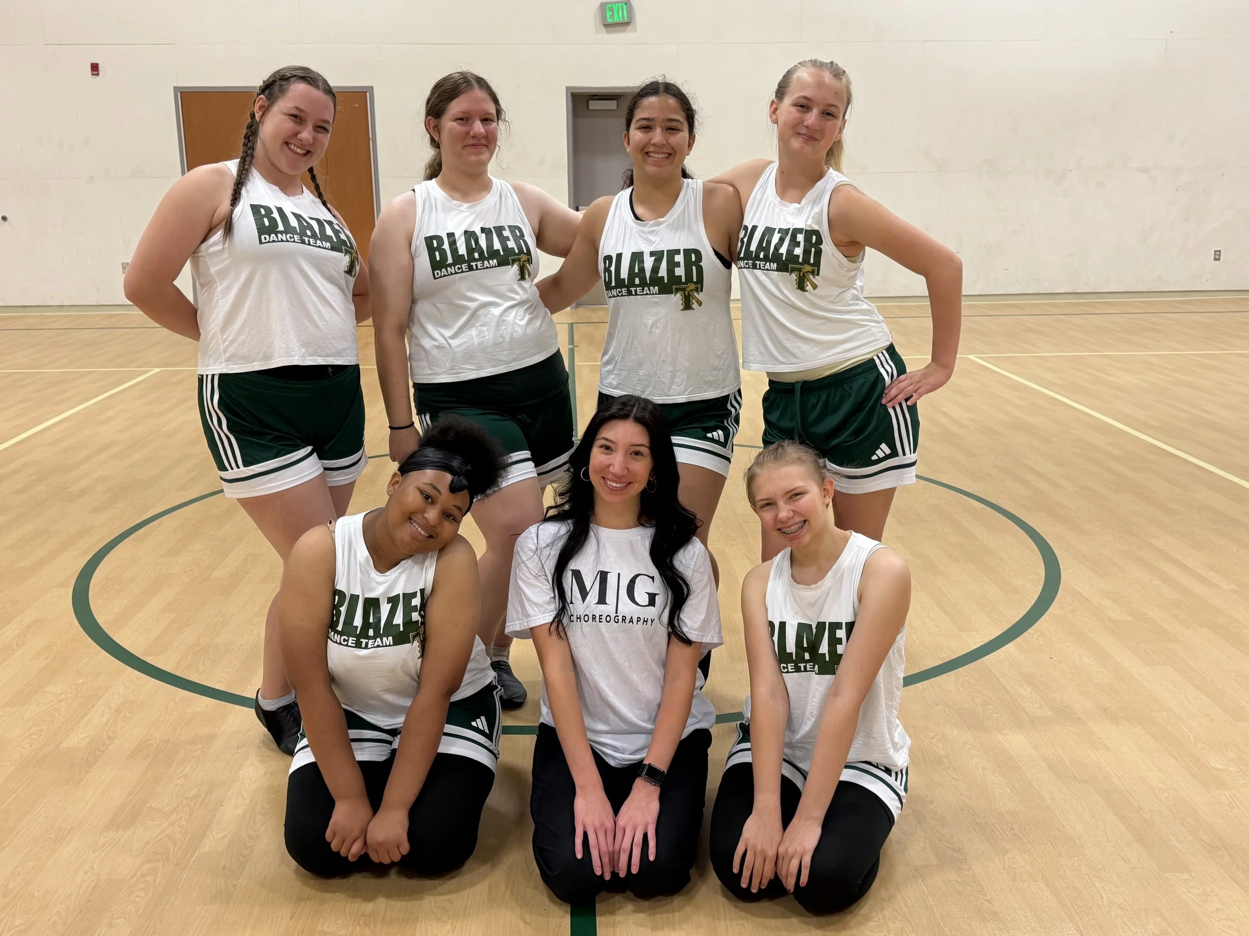 Group of young women in athletic uniforms posing indoors on a basketball court, smiling for the camera.