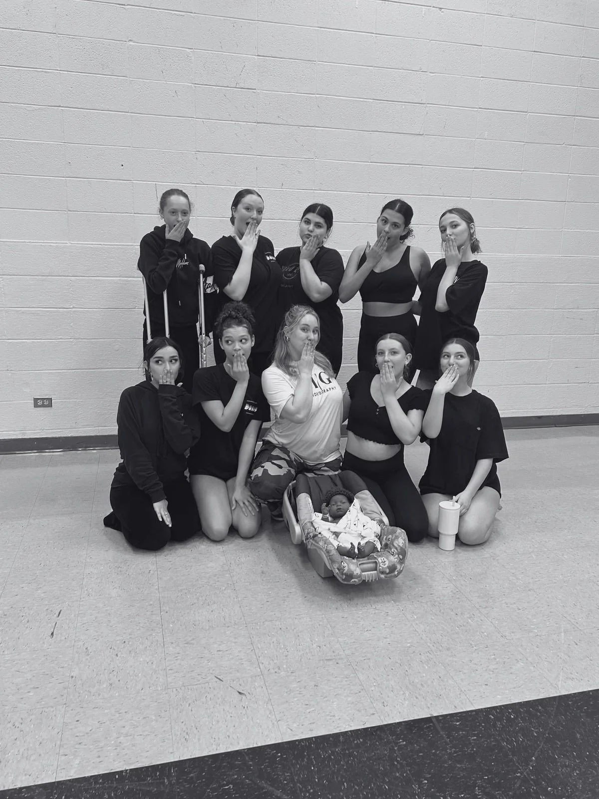 Group of eleven young women, some kneeling and some standing, all covering their mouths with one hand in a gesture of surprise or secrecy, posing in a gymnasium with a brick wall background. There is a baby in a car seat and a woman in the middle in 