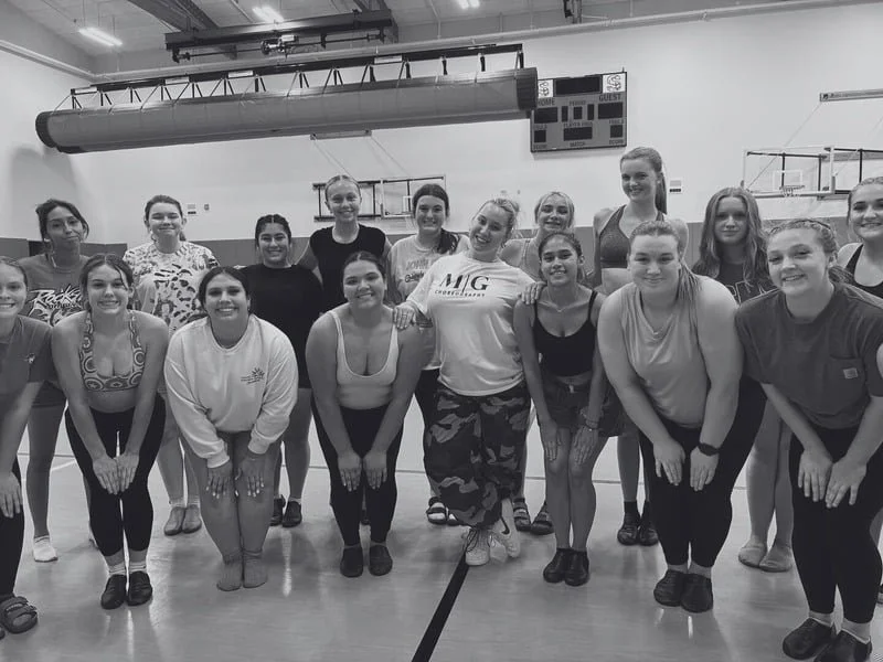 Group of young women posing in a gymnasium, some with their hands on knees, smiling at camera.