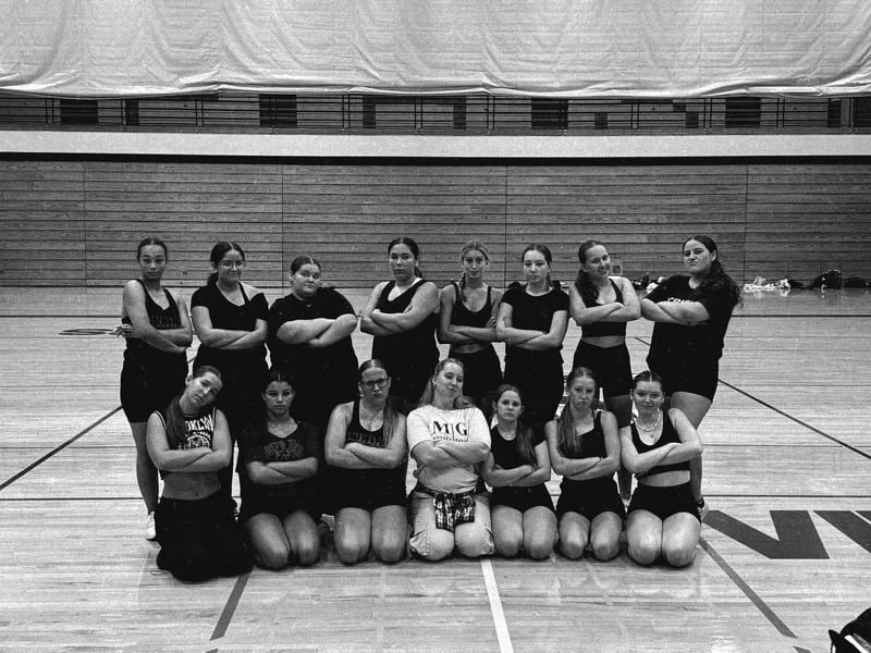 A group of 14 young female volleyball players in athletic wear posing with arms crossed in an indoor gymnasium.
