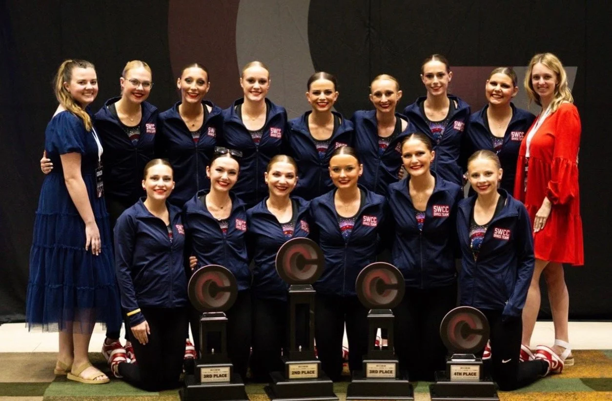 A group of young women in matching navy jackets with 'SWCC' logos, posing with trophies at an indoor event. They are smiling for the photo, with some kneeling in front and others standing behind. One woman on the far right is wearing a red dress.