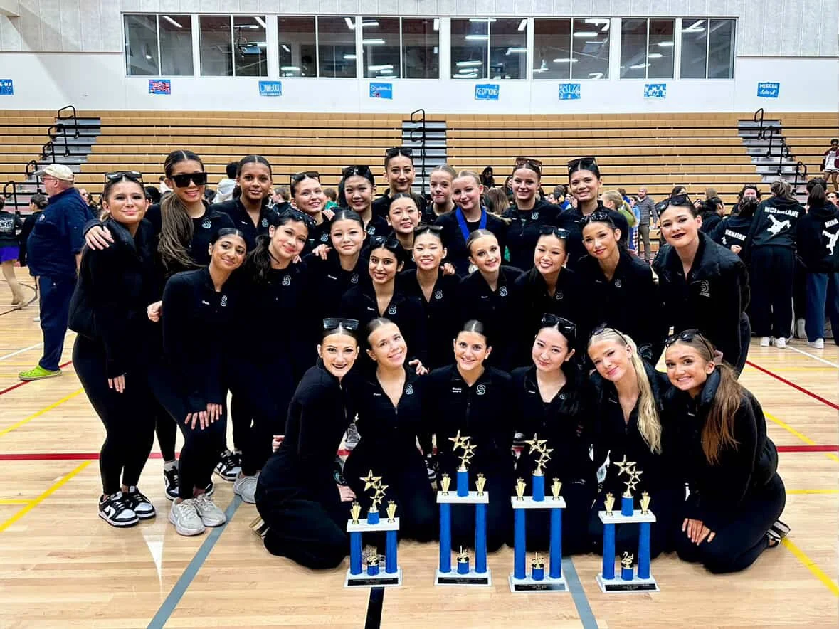 A group of women dressed in black athletic outfits posing with trophies in a gymnasium.