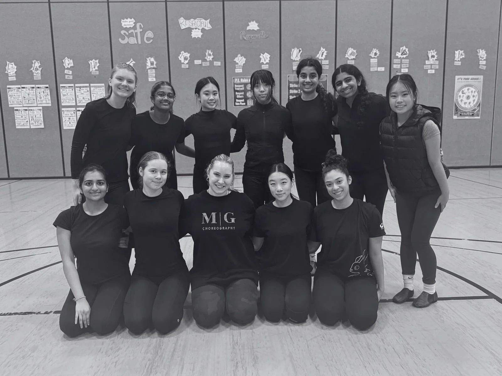 A group of twelve young women in black clothing, posed in a basketball gymnasium for a photo. The background features posters on the wall and the gym floor with court lines.