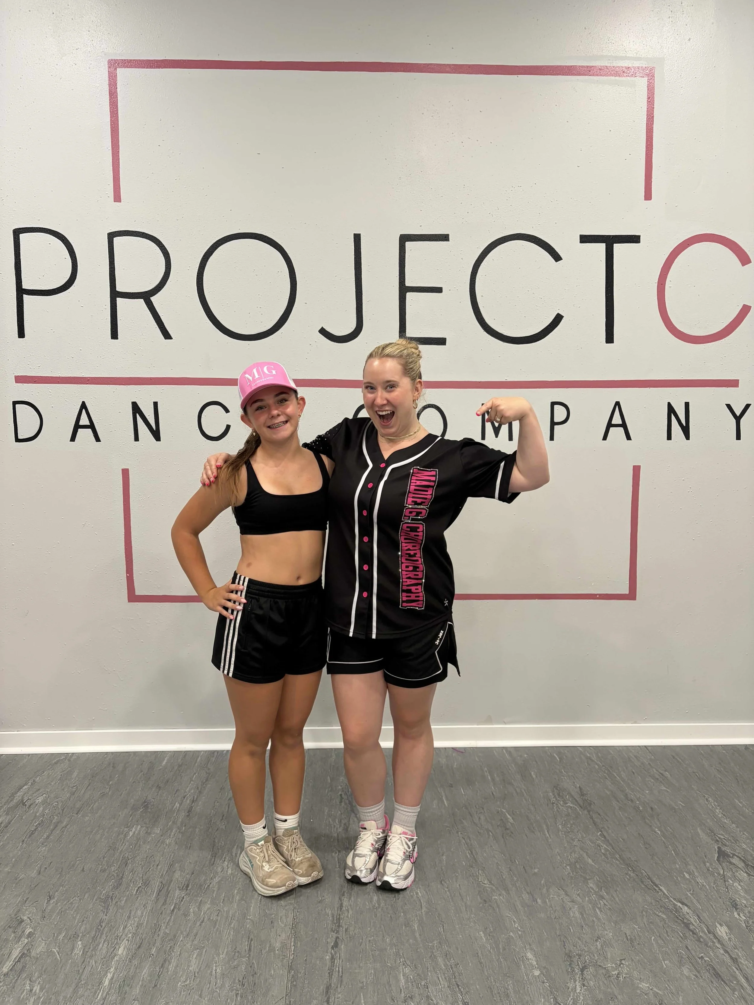Two young women in athletic clothing stand in front of a wall with a dance studio logo. One woman is wearing a pink cap, black crop top, black shorts, and beige sneakers. The other woman has blonde hair tied back, wearing a black and pink dance team 