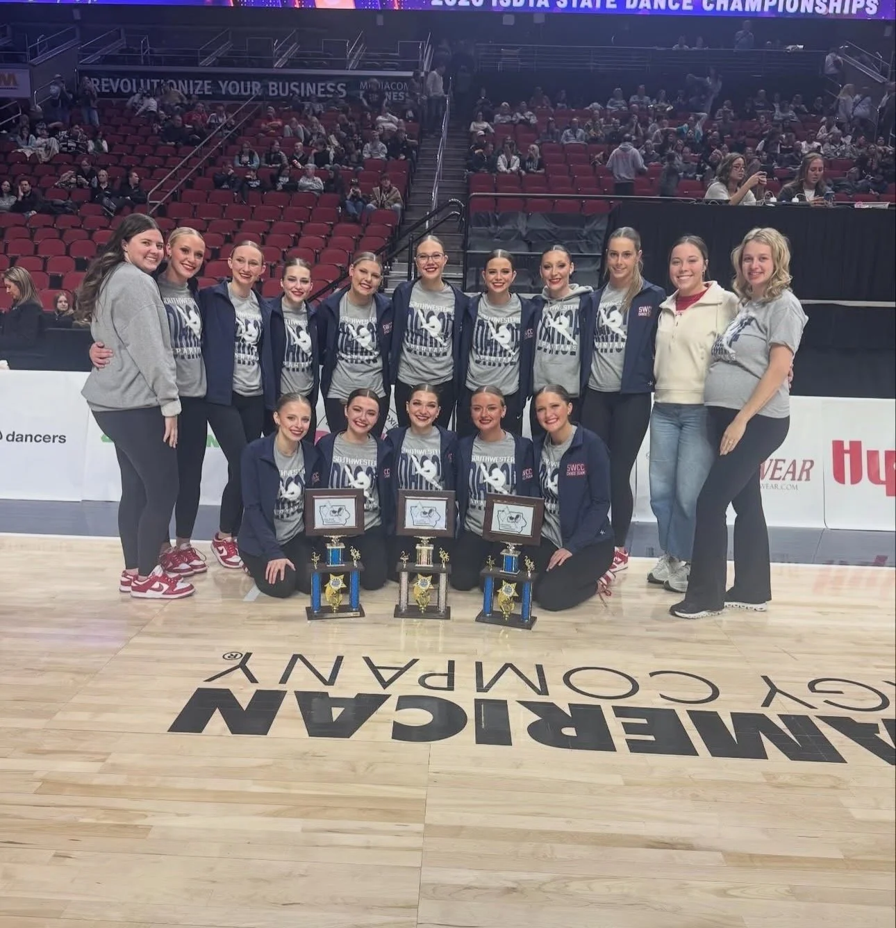 A group of young women, likely a dance team, standing on a basketball court with trophies and plaques in front of them. They are wearing matching gray shirts and navy jackets with the words SWIC and a logo. Behind them are empty red seats and a crowd
