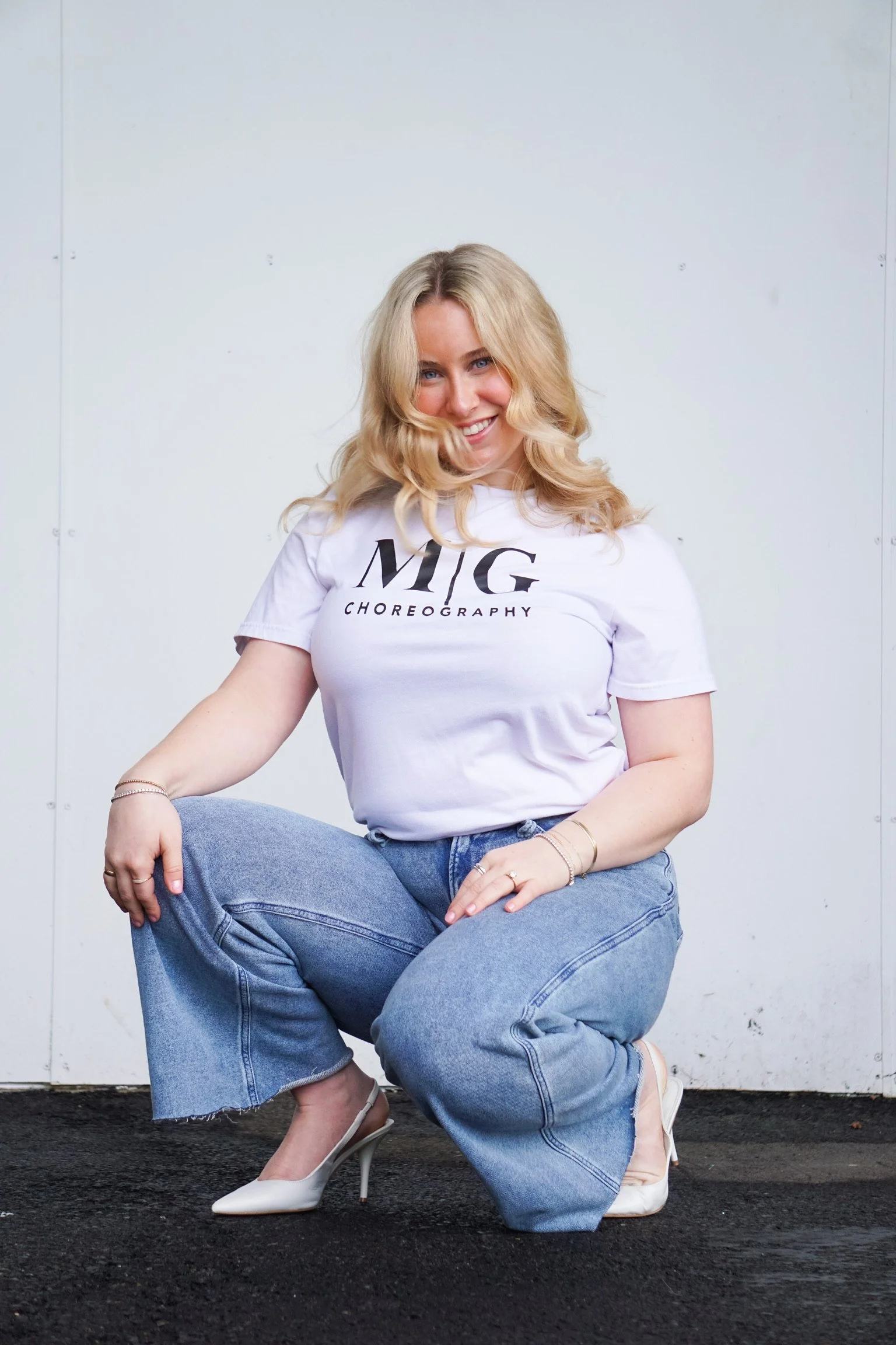 A smiling blonde woman wearing a white t-shirt with black text that says 'MIG CHOREOGRAPHY,' blue jeans, and white high heels, sitting on her heels on a black asphalt ground in front of a plain white wall.