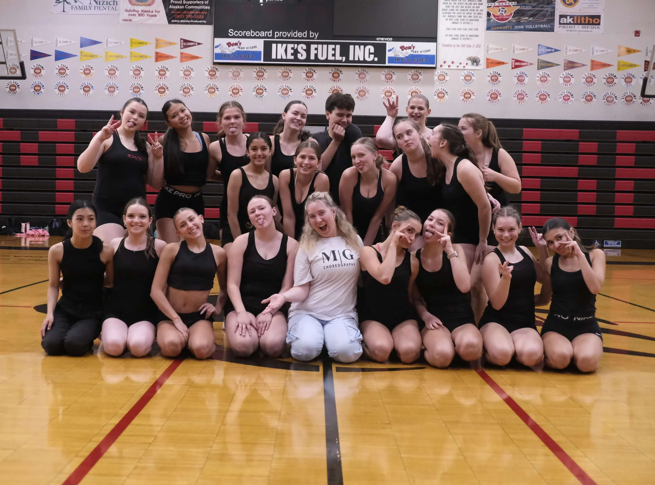 Group of young girls in black athletic outfits posing for a photo in a gymnasium. Some are kneeling in the front row, others standing behind them. The background features a bulletin board with various banners and awards.