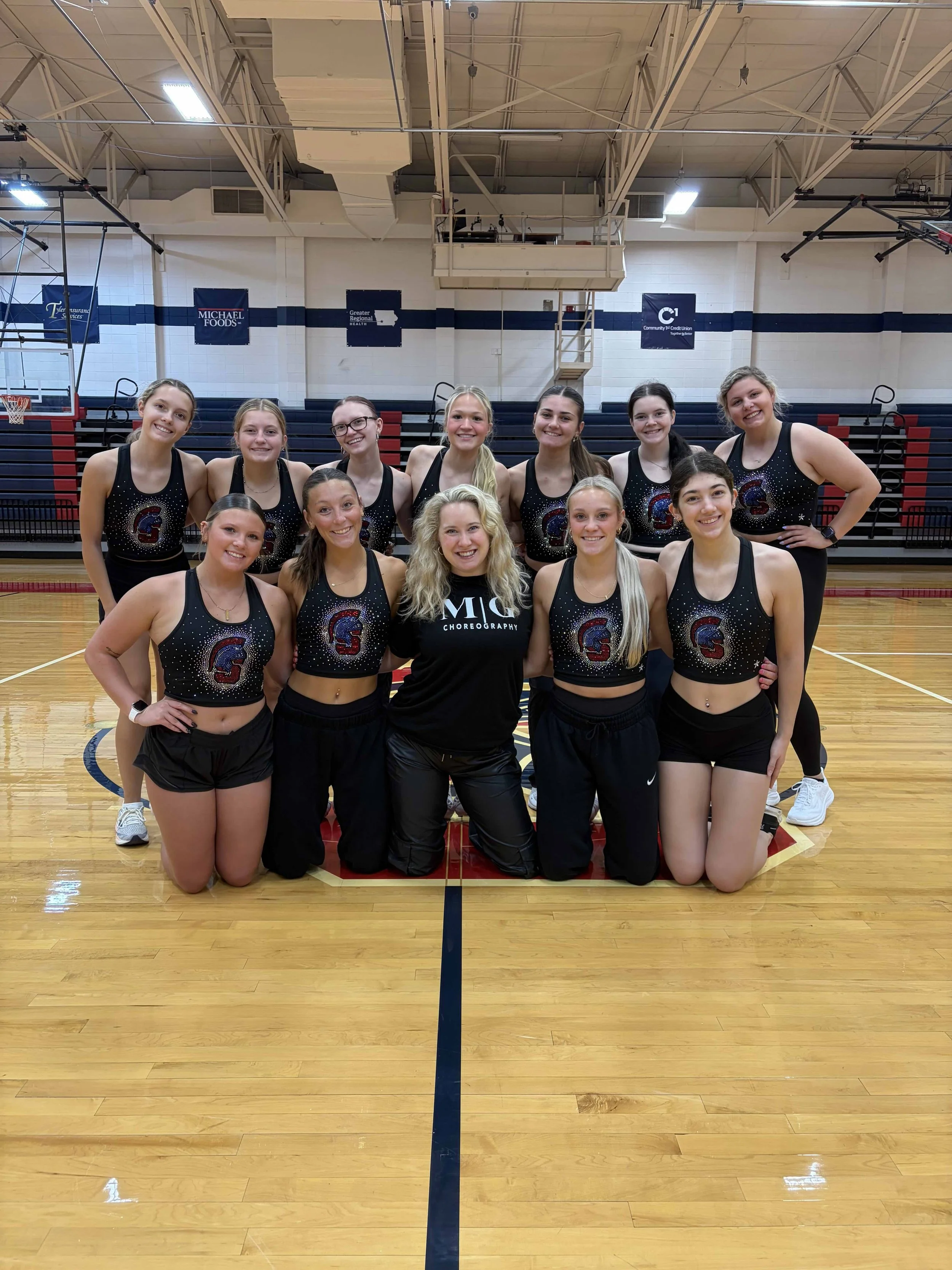 A group of ten young women wearing matching black cheerleading uniforms with a multicolored 'G' logo, posing in a gymnasium with a basketball court, bleachers, and banners in the background.