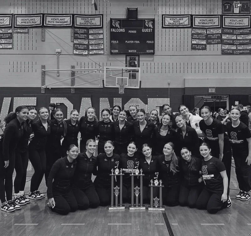 Group of young women athletes in a gymnasium, posing with trophies, wearing sports outfits and jackets with an 'S' logo.
