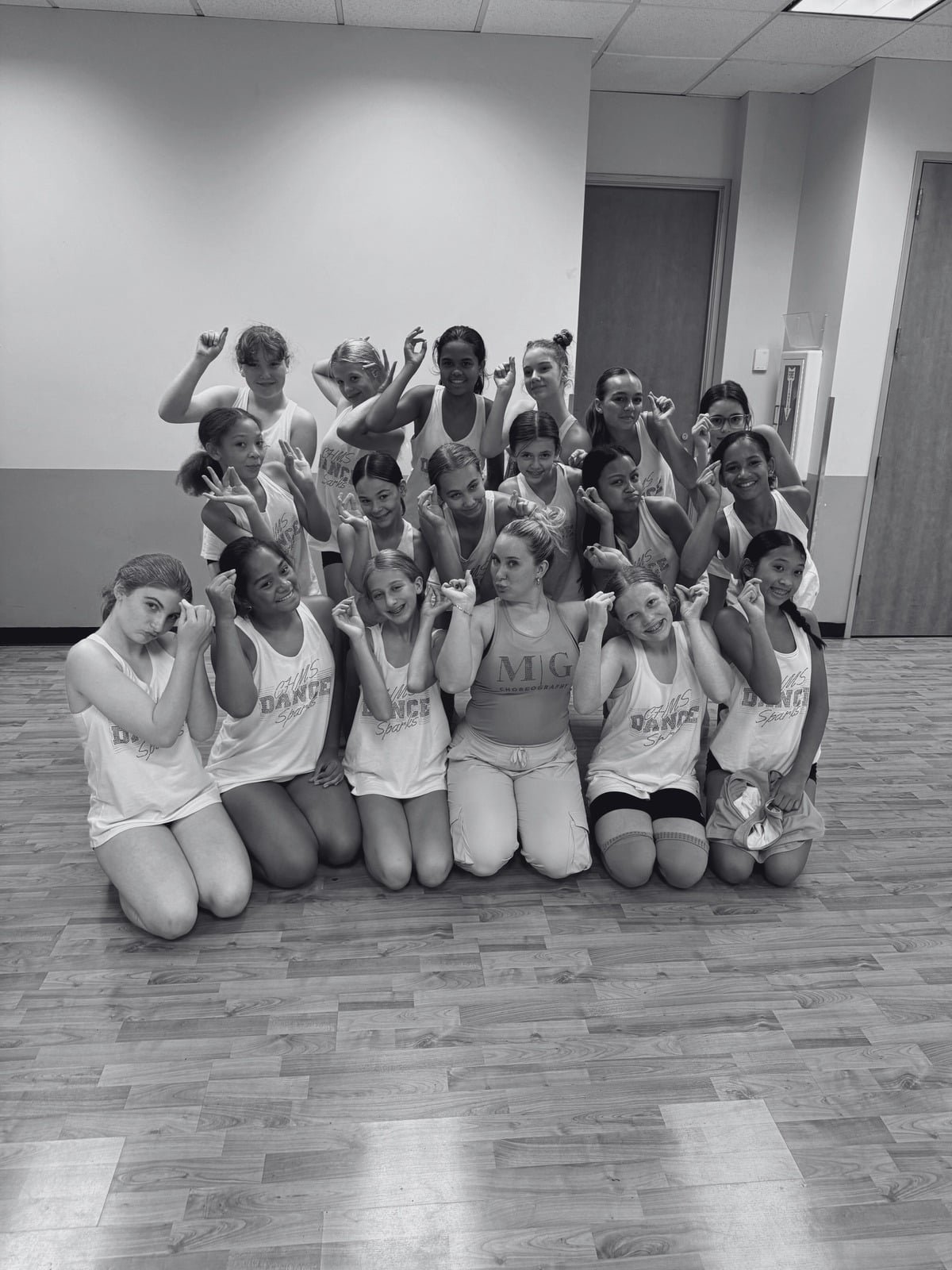 A group of young girls in dance costumes and one adult instructor posing together in a dance studio, with many of them making pointing or peace signs with their hands.