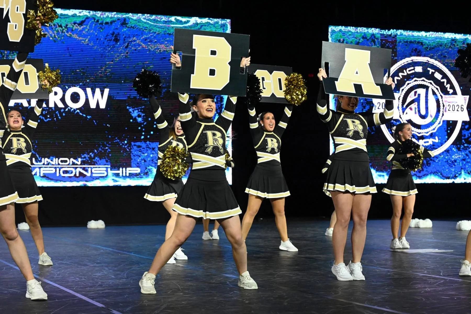 Cheerleaders in black and gold uniforms perform a cheer routine on stage during a UCA cheerleading championship, holding signs that spell out 'B A C K' against a digital backdrop with the words 'Union Championship'.