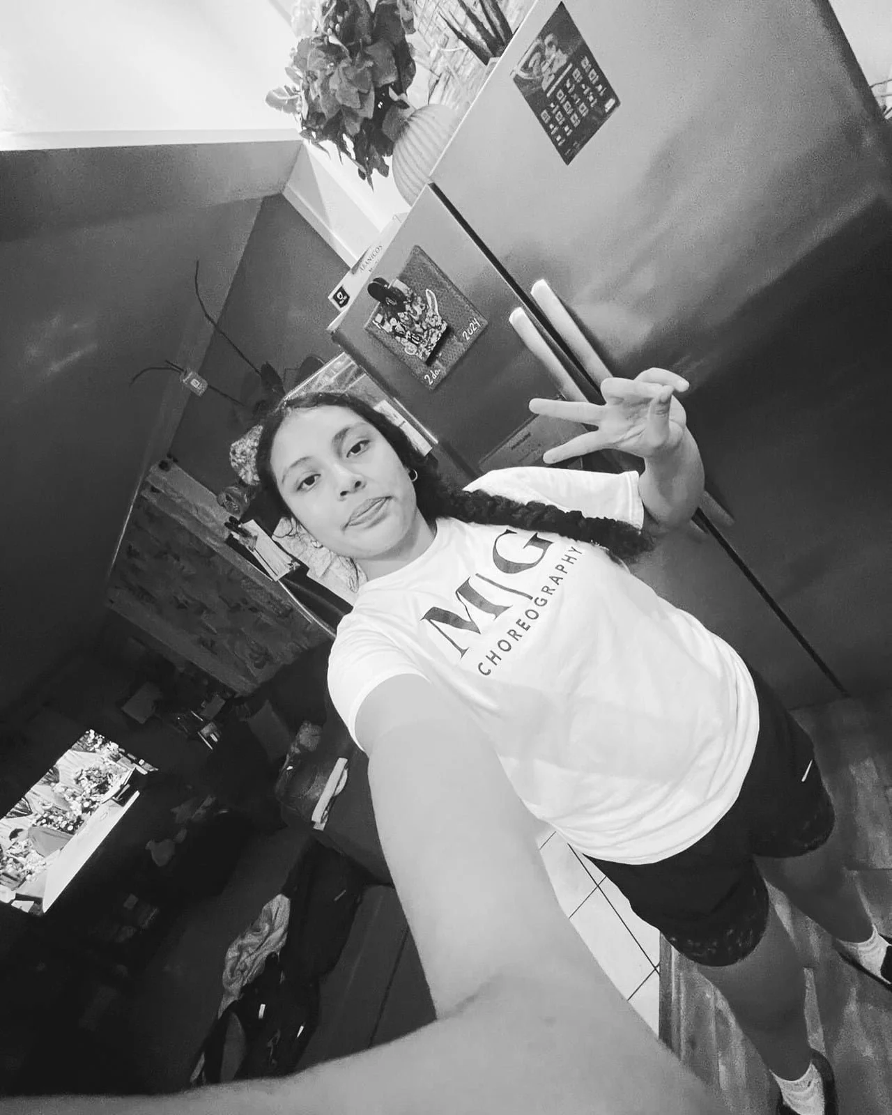 A young woman taking a selfie in a kitchen, making a peace sign with her left hand, wearing a t-shirt that says 'VIC CHOREOGRAPHY', with a fridge, a TV, and kitchen cabinets in the background.