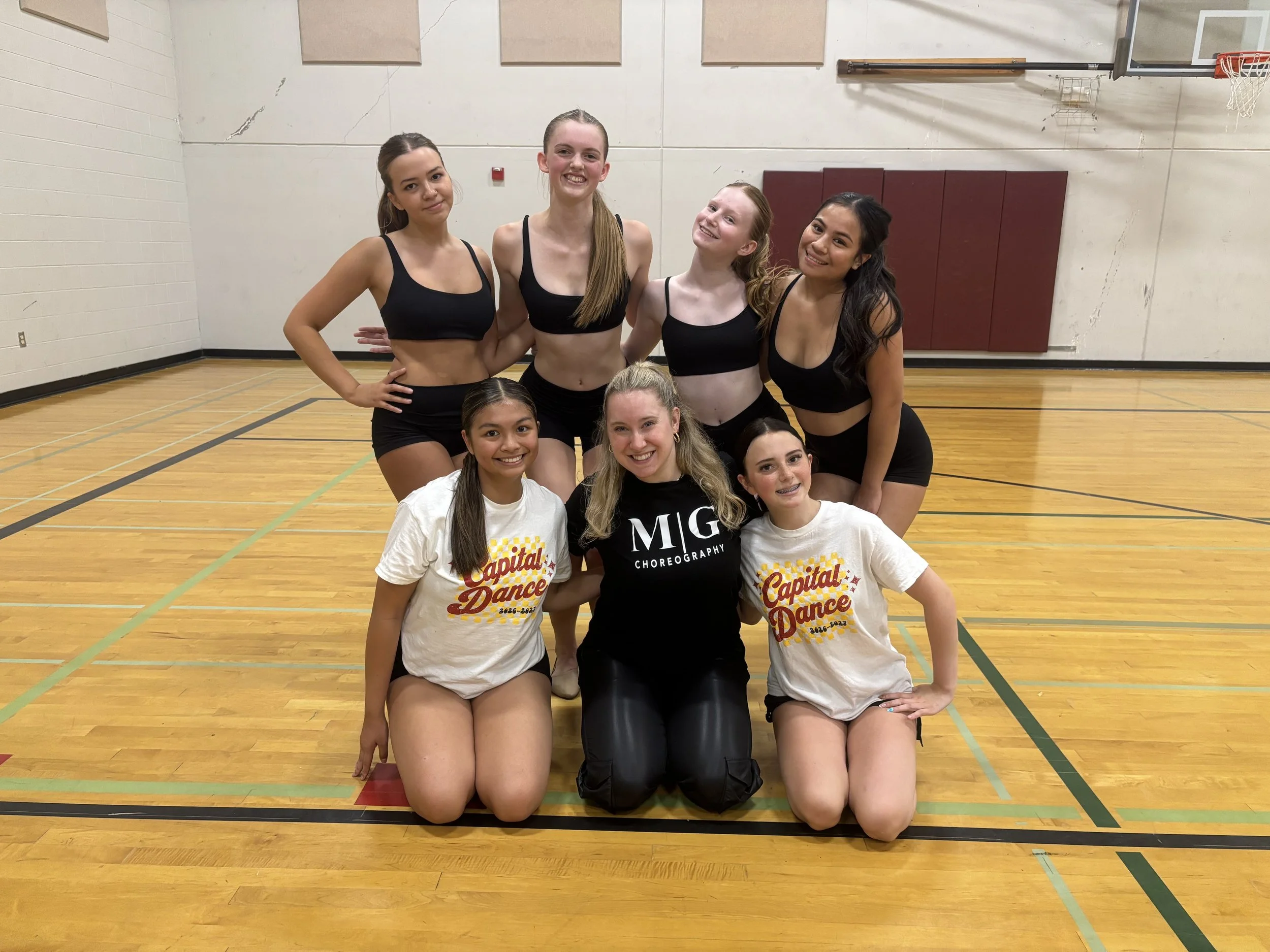 Group of six young women in a gymnasium, some kneeling and some standing, smiling at the camera. They are dressed in athletic wear and have their hair tied back. The gym has a hardwood floor, basketball hoop, padded wall, and some wall-mounted equipm