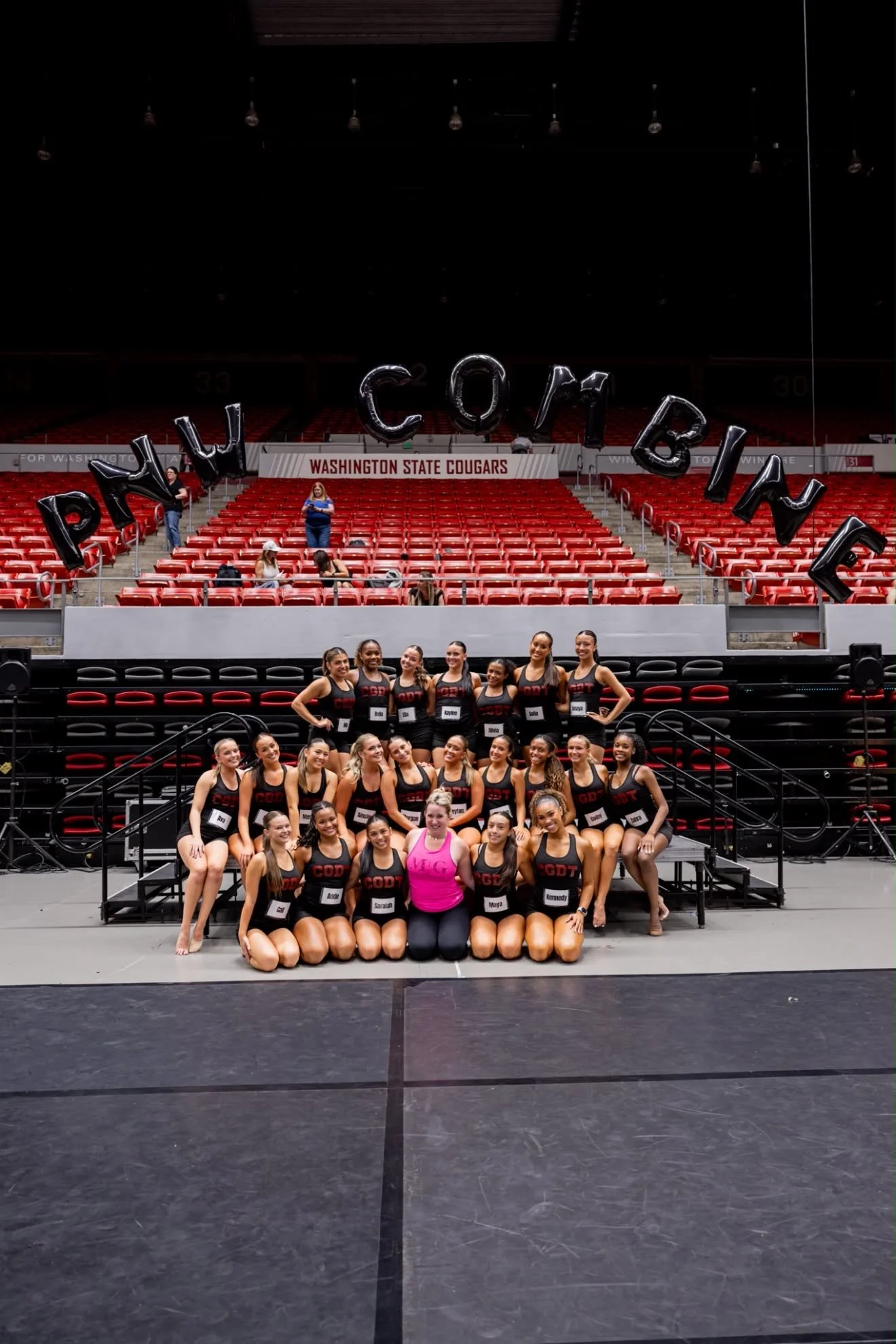 Group of young female gymnasts in black and red uniforms posing on a stage with a woman in a pink shirt, in an indoor arena with red seats and a banner reading 'WASHINGTON STATE COUGARS' in the background.