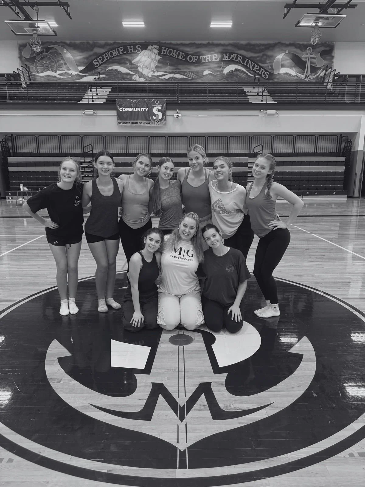 Group of young female cheerleaders in athletic clothing, smiling, posing on a basketball court, with a Sehome High School banner and mural in the background.