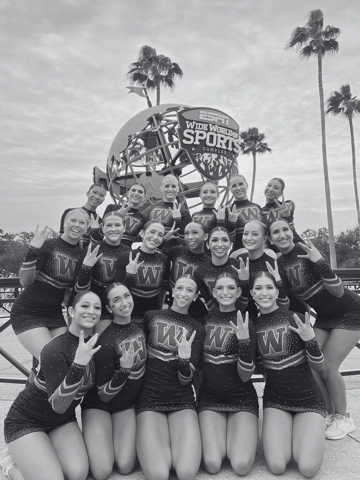 A group of cheerleaders in matching uniforms, posing together outdoors in front of a large globe sculpture with a sign that reads 'ESPN Wide World of Sports Complex.' The cheerleaders are smiling, making peace and victory signs, and kneeling in front