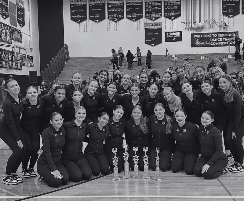 Group of young female athletes in matching athletic uniforms posing in a gymnasium with four trophies.