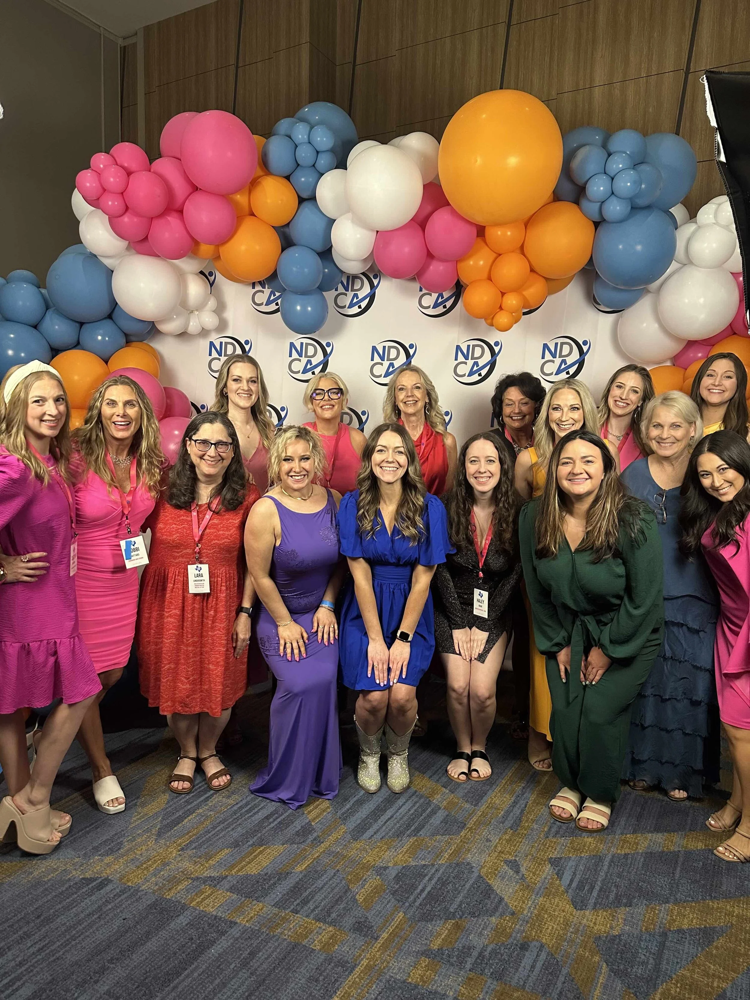 Group of women smiling, standing in front of a backdrop with the 'NDCA' logo, surrounded by colorful balloons in pink, blue, orange, and white.