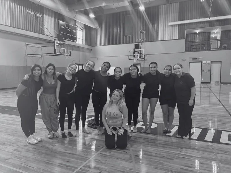 Group of ten women in sportswear standing together in an indoor basketball court, some with arms around each other, with a woman kneeling in front.