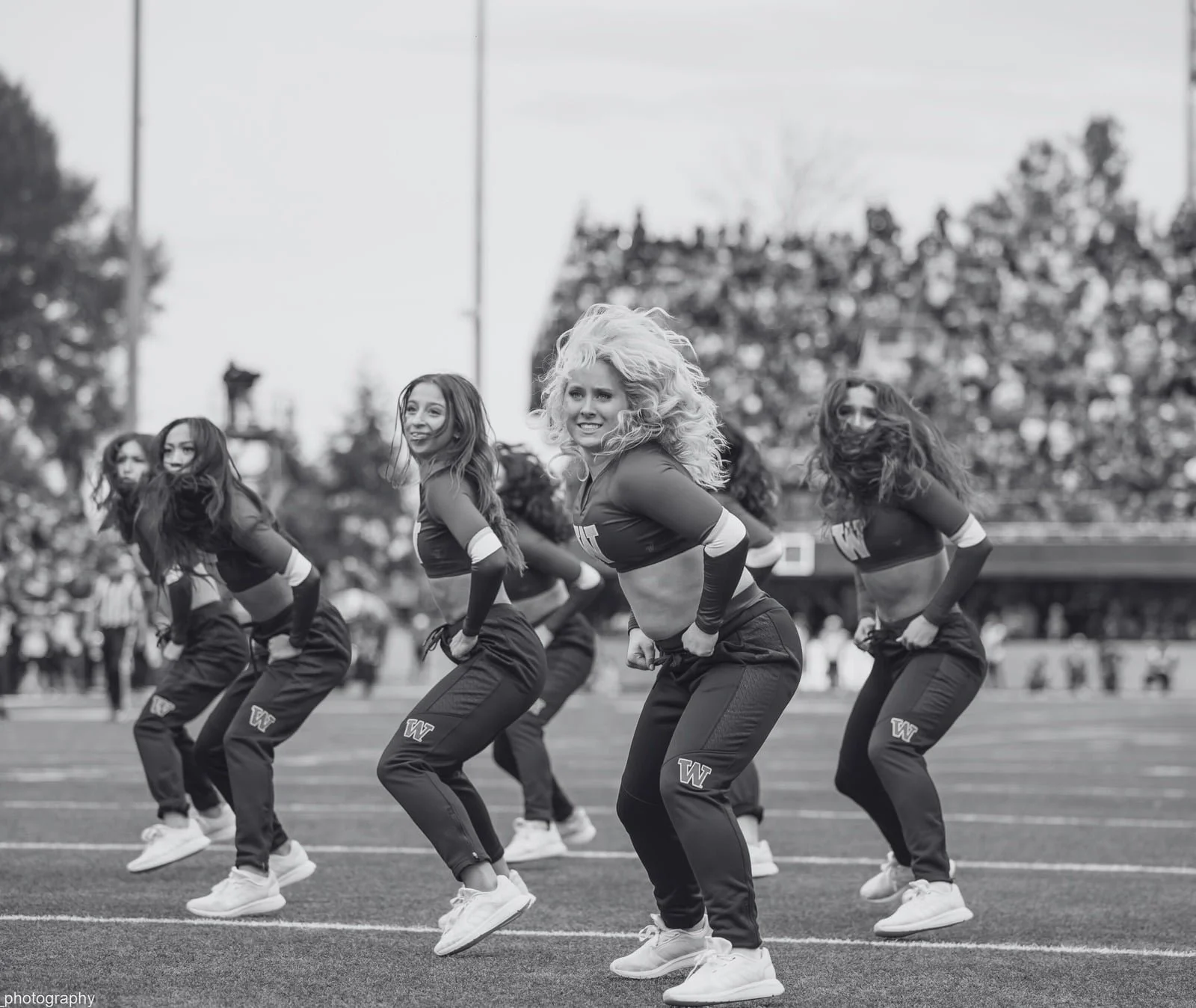 A group of cheerleaders performing a dance routine on a football field, wearing coordinated outfits with a 'W' logo, with spectators and trees in the background.