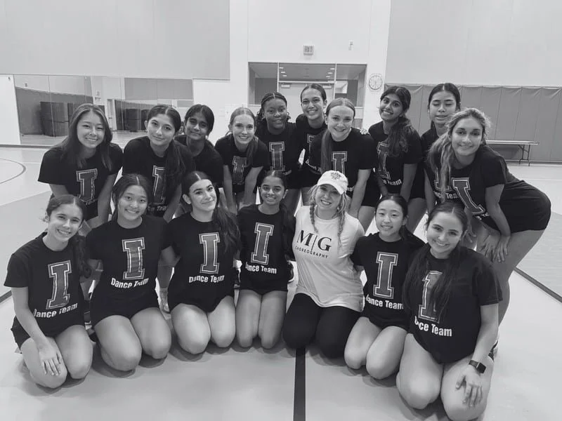 Group of young female dancers in black T-shirts with 'L Dance Team' lettering, posing in a gym, with one woman in a white T-shirt and cap.
