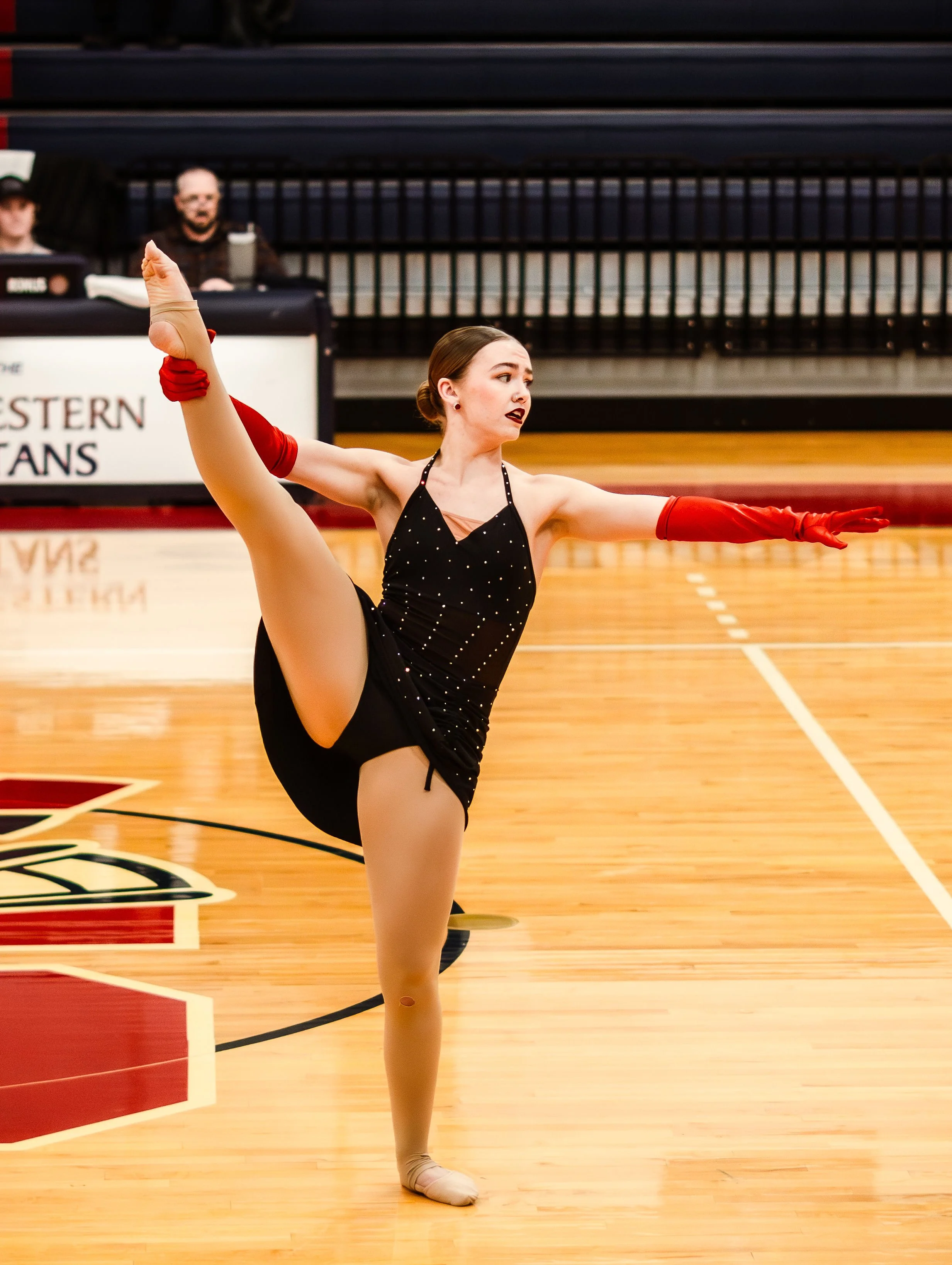 A young female ballet dancer in a black dress with small sparkles performing a high leg extension on a wooden gymnasium floor, with an audience and judges in the background.