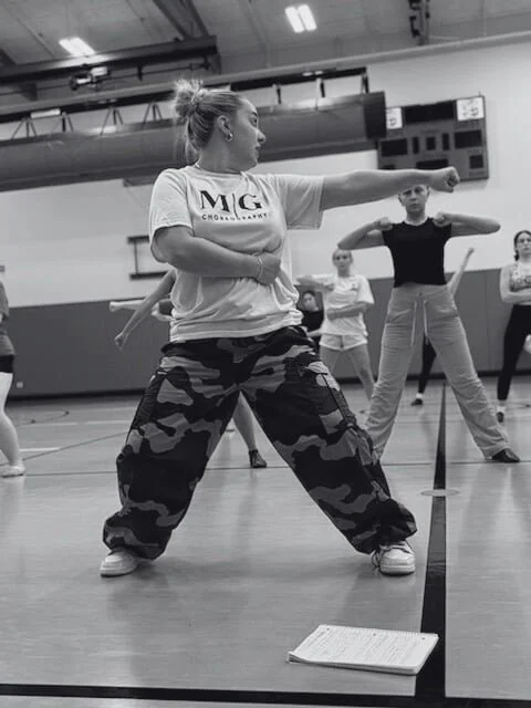 A woman in a t-shirt and camouflage sweatpants demonstrating a martial arts pose during a class or rehearsal in a gymnasium, with other women watching her.