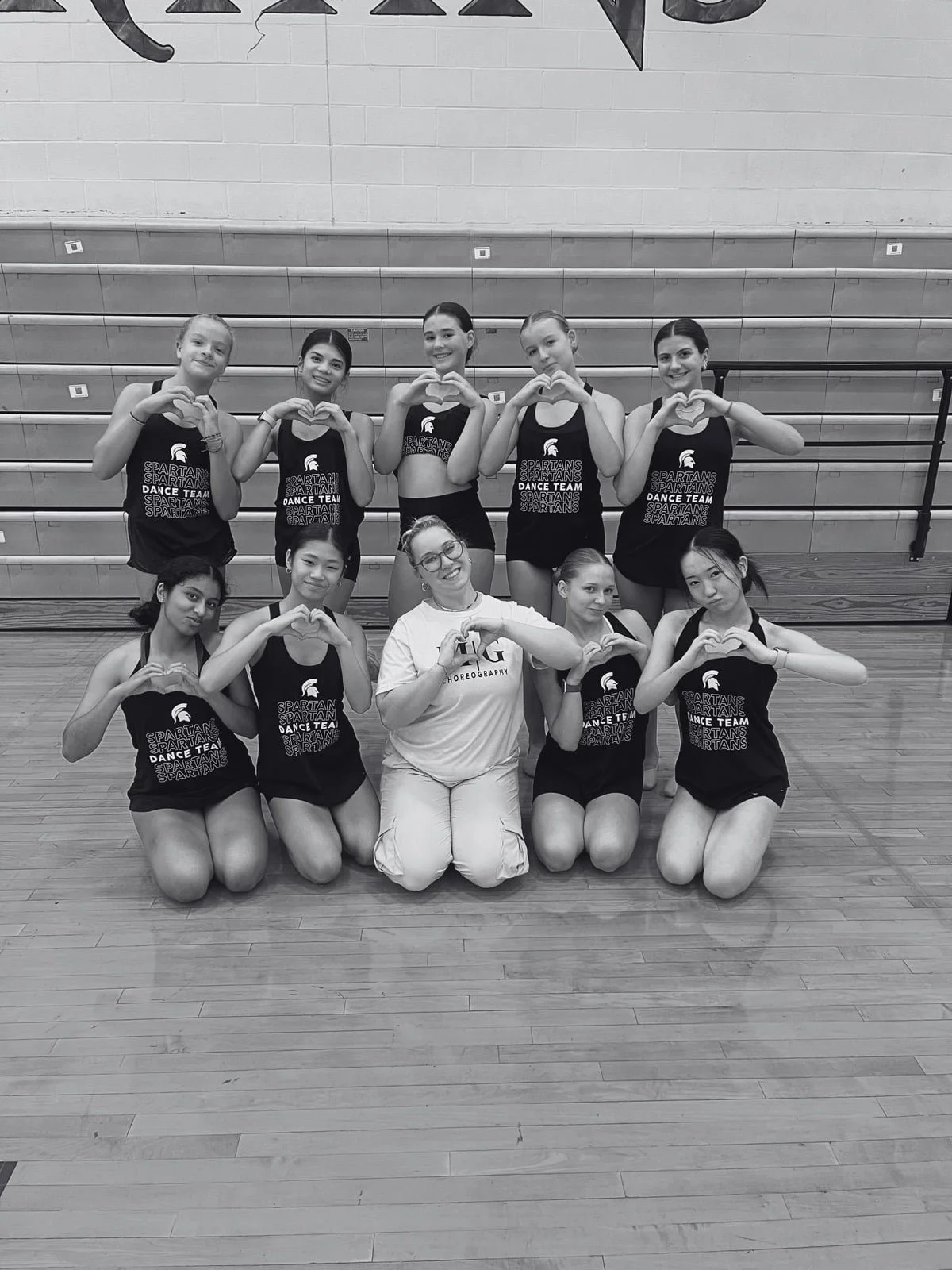 A group of female dancers in athletic outfits and a coach in a gym, all making heart signs with their hands. The girls are wearing tank tops with the text 'Spartans Dance Team,' and the coach is in a white shirt with the word 'Choreography' on it.