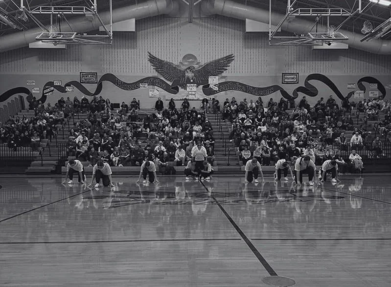 Group of people performing push-ups on a basketball court with an audience seated in the bleachers and a large eagle mural on the wall behind.