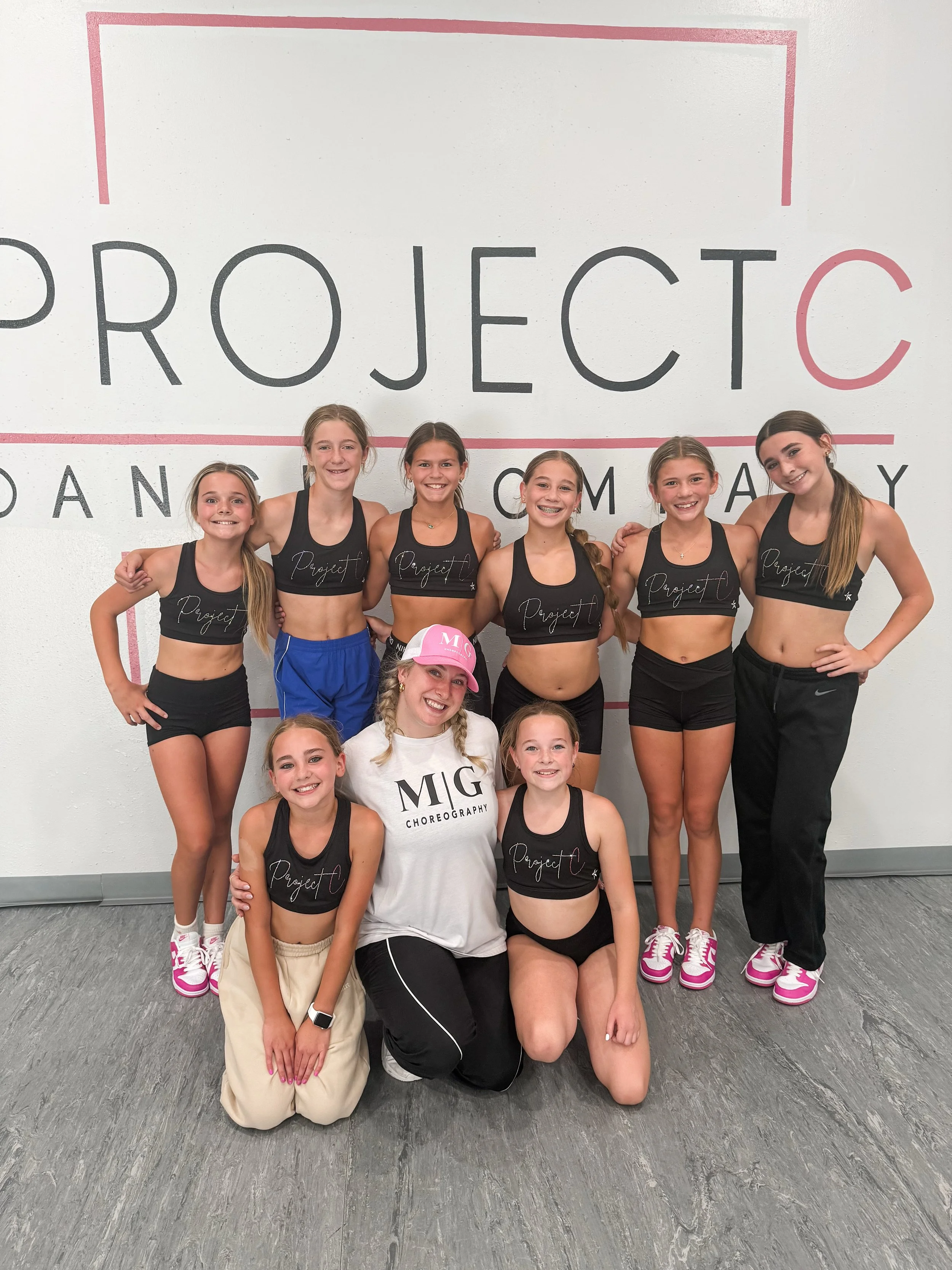 A group of nine cheerleaders, mostly young girls with one adult woman, smiling and posing in front of a wall with the words 'PROJECT' and 'CHOREOGRAPHY' written on it, wearing black sports bras with 'Project' written on them and athletic shorts or pa