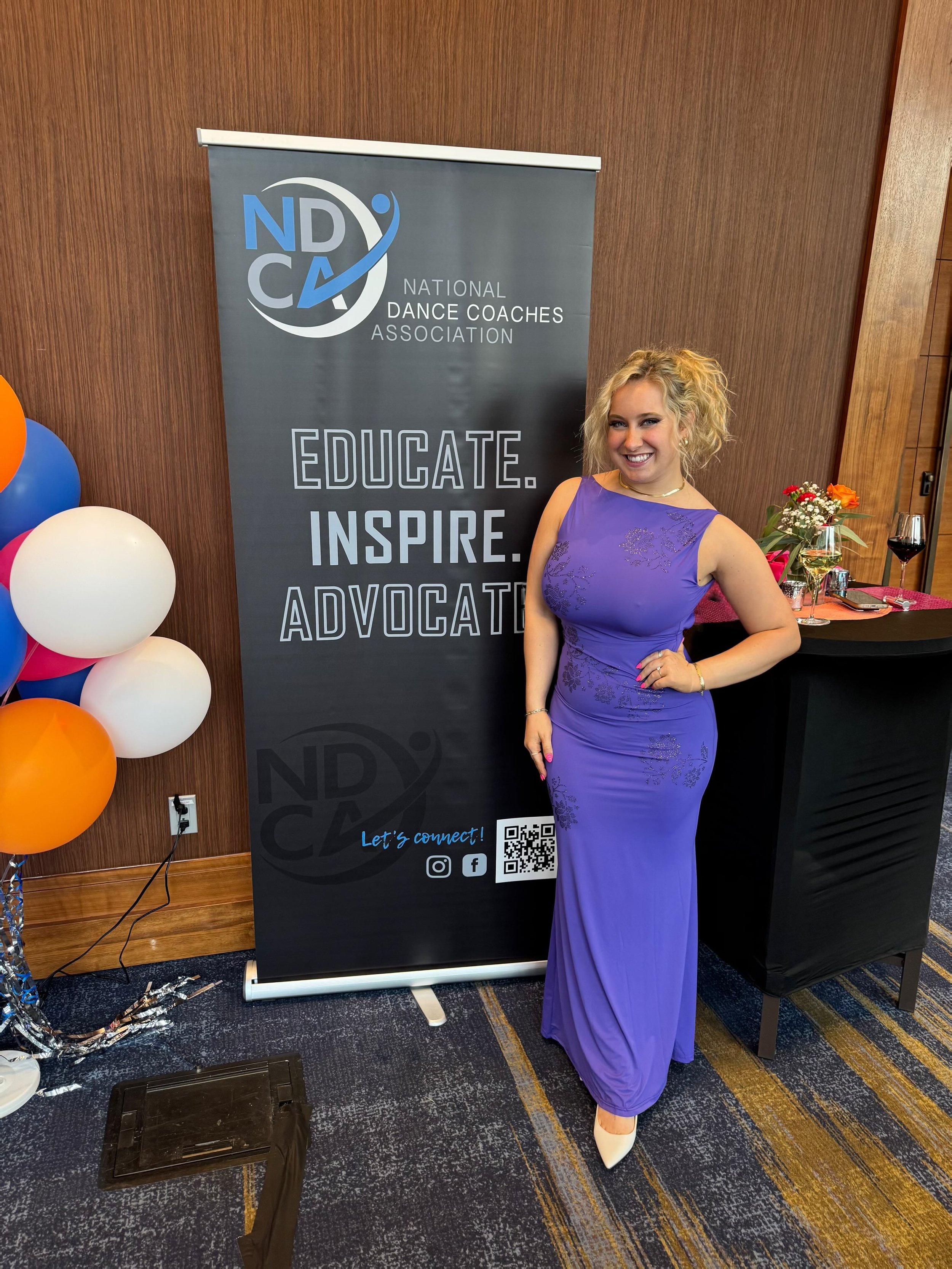 A woman in a purple evening gown standing beside a banner for the National Dance Coaches Association, smiling at the camera. The banner features the NDCA logo and the words 'Educate, Inspire, Advocate.' There are colorful balloons and a table with fl