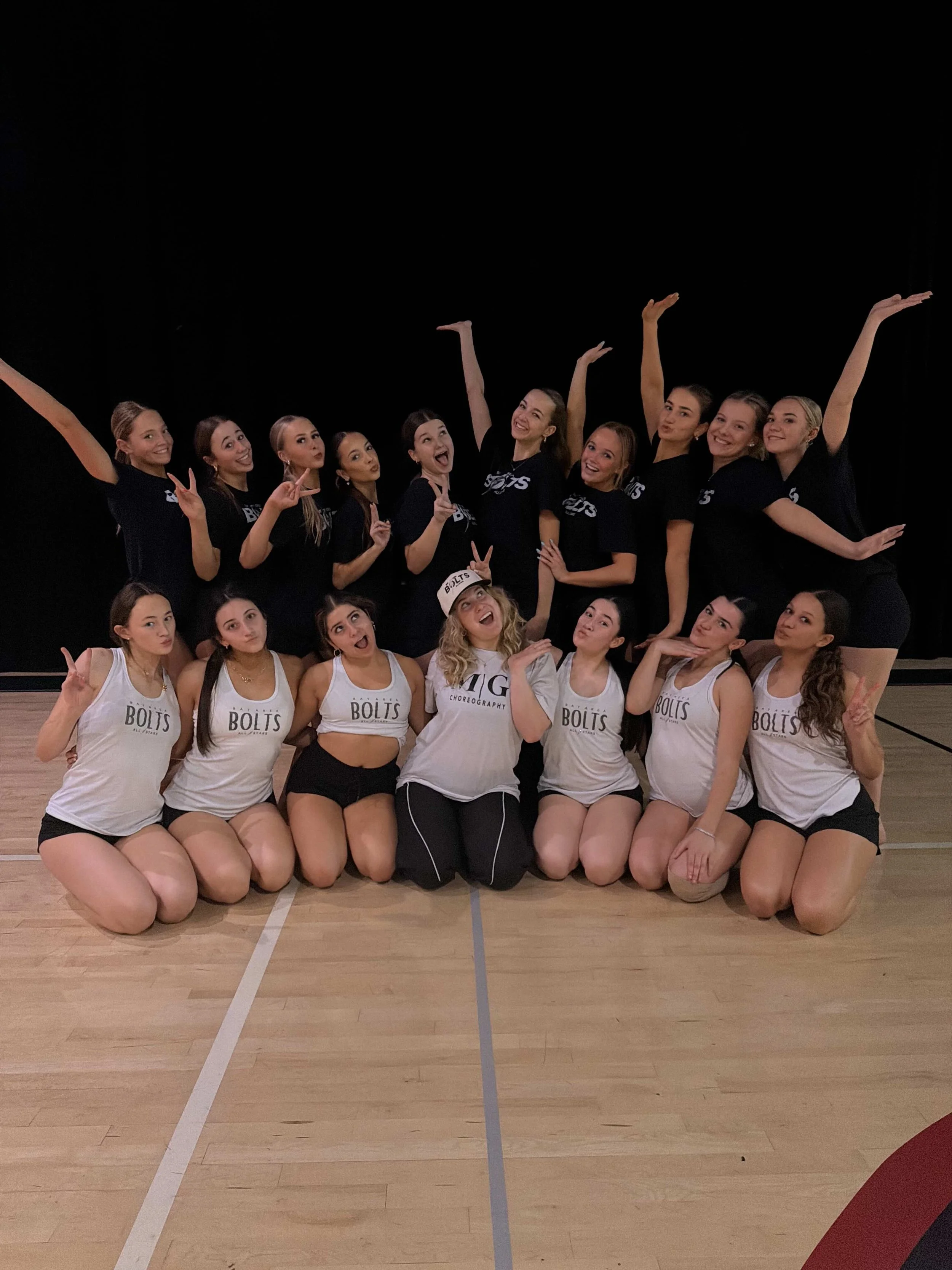 Group of young women in cheerleading uniforms, some standing and some kneeling, posing on a wooden gymnasium floor with a black background.