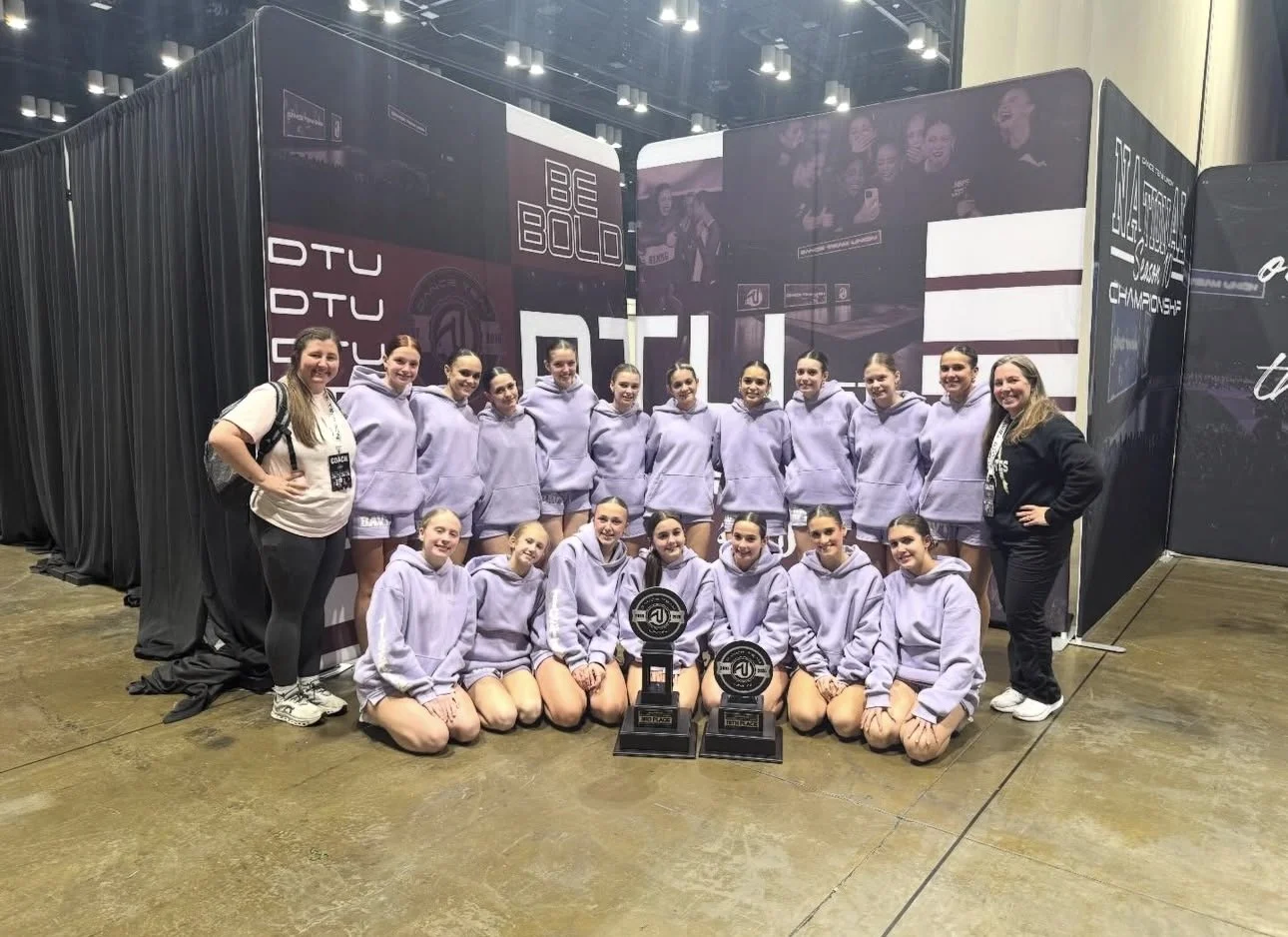 A group of female athletes in gray hoodies posing with trophies at an indoor sports event.