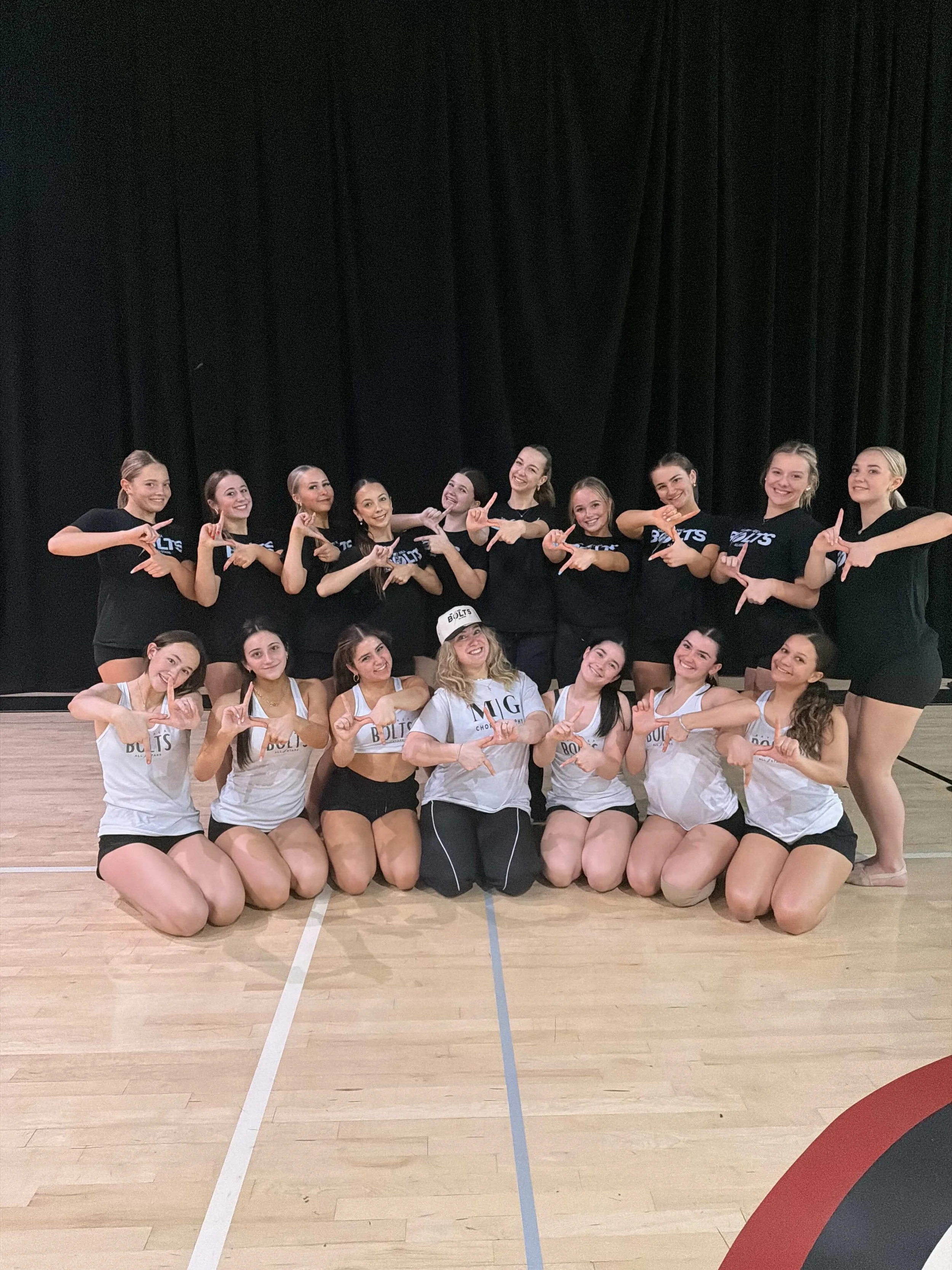 Group of young women in athletic clothing posing for a photo in an indoor gym, smiling and making hand gestures.