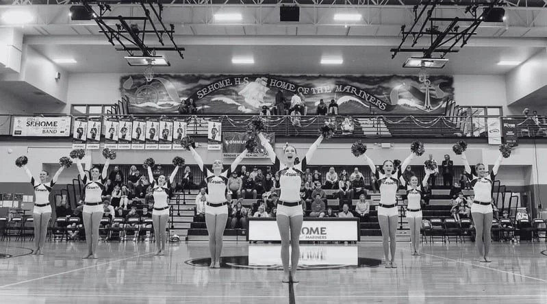 Cheerleaders performing a routine with pom-poms in a gymnasium, with spectators in the background.