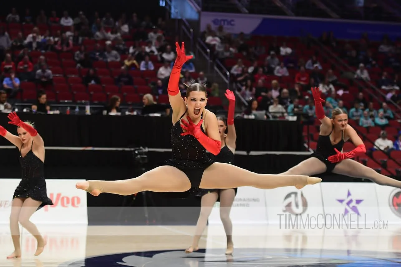 A group of female gymnasts performing a synchronized jump during a routine, with audience members watching from the stands.