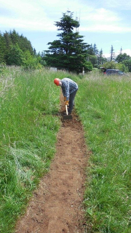 A Cape Horn Conservancy volunteer with an orange hard hat digging a trail in a grassy field, with trees and a car in the background.