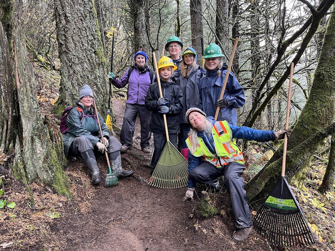 Group of seven people wearing rain gear and hard hats, holding rakes, in a forested area, likely doing trail maintenance.
