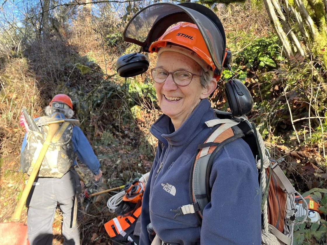 Smiling woman wearing an orange helmet with a headset, glasses, and outdoor gear, standing in a forested area with light filtering through the trees.