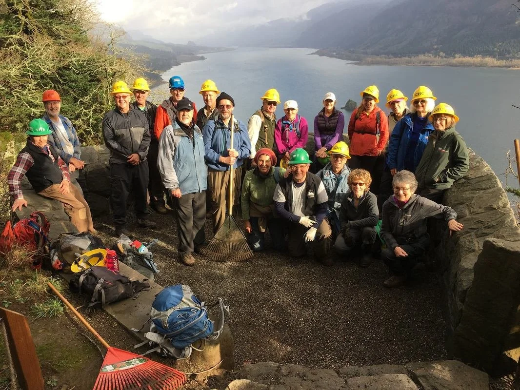 Group of Cape Horn Conservancy volunteers wearing safety helmets and outdoor clothing, gathered on a rocky overlook by the Columbia River Gorge and mountains, with tools and backpacks in the foreground.
