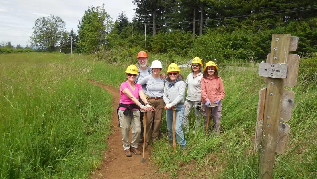 Group of six, Cape Horn Trail volunteers wearing yellow safety helmets, standing on a grassy trail in a forested area during daytime, with a wooden Cape Horn Trail sign nearby.