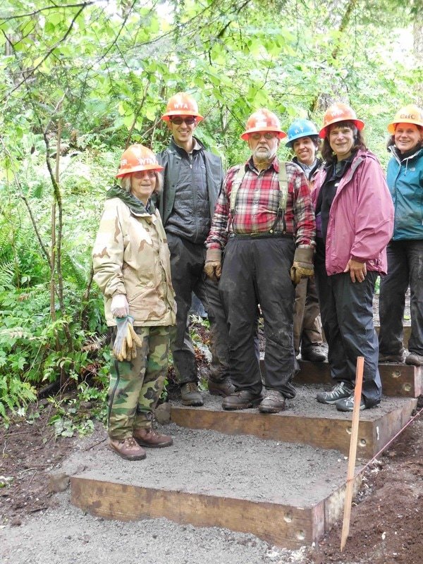 Group of Cape Horn Conservancy volunteers wearing safety helmets and outdoor clothing standing on a partially constructed wooden staircase in a forested area.