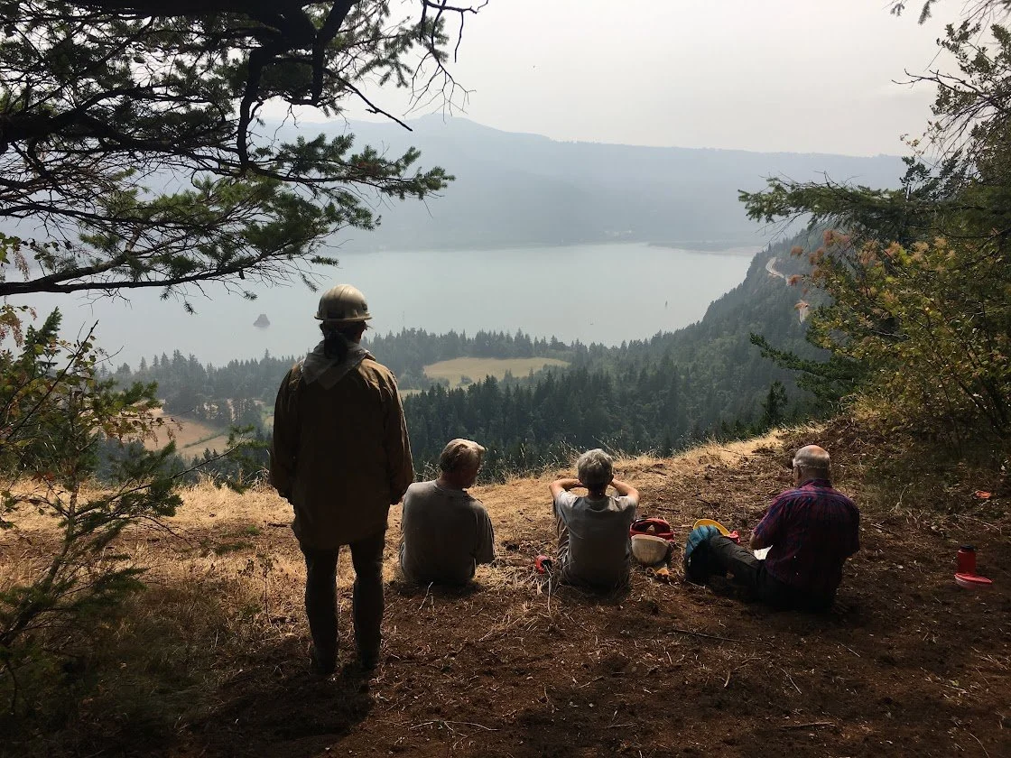 Four Cape Horn Conservancy volunteers sitting on a grassy hillside overlooking the Columbia River Gorge and forested mountains, with one person standing nearby in a cap and jacket.