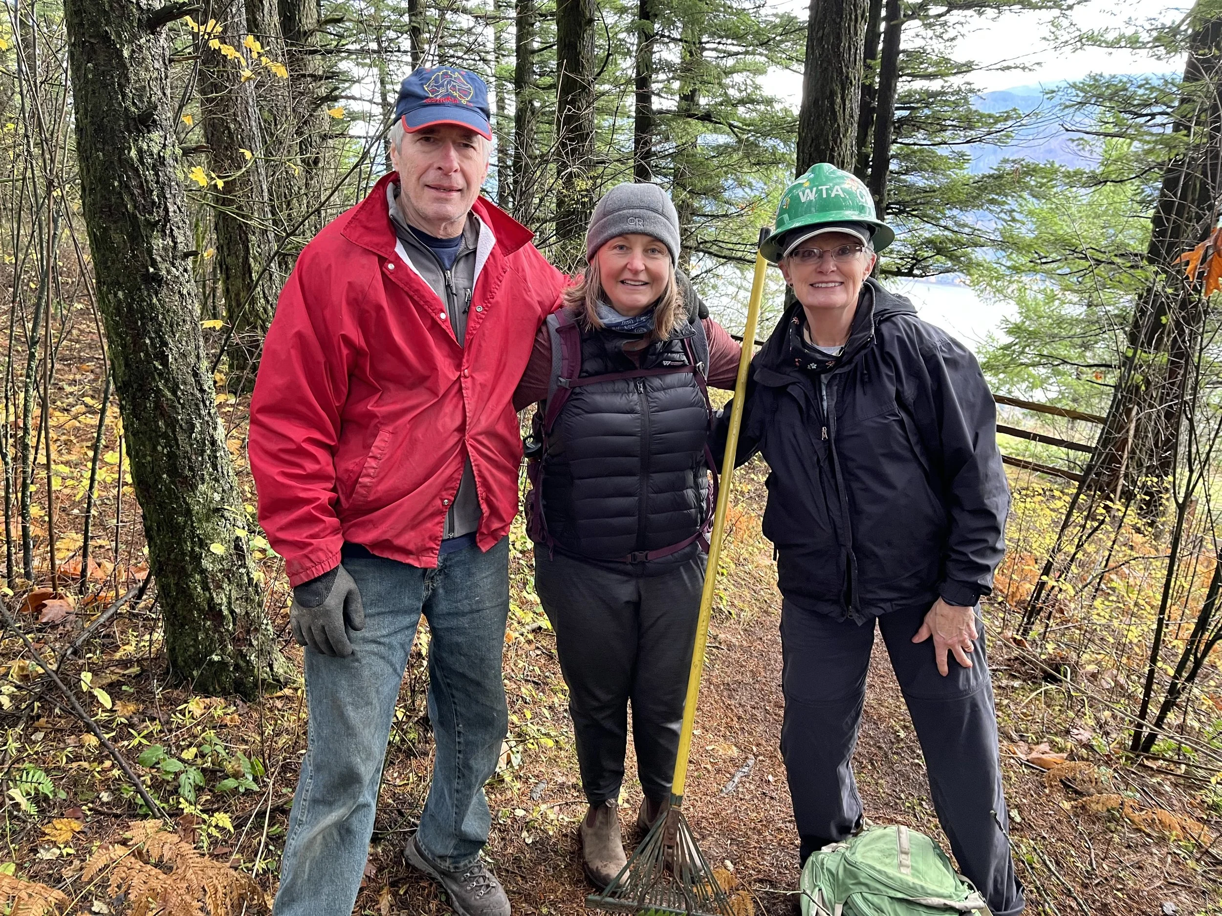 Three Cape Horn Conservancy volunteers in hiking gear standing in a wooded area with trees and fallen leaves, posing for a photo.