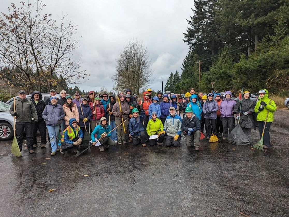 Group of volunteers for the annual Rake a thon in rain gear holding rakes outdoors on a cloudy day, near parked cars and trees.