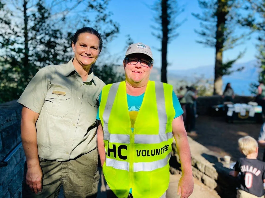 Two women, one in a volunteer vest and the other in a uniform, smiling outdoors with trees in the background.